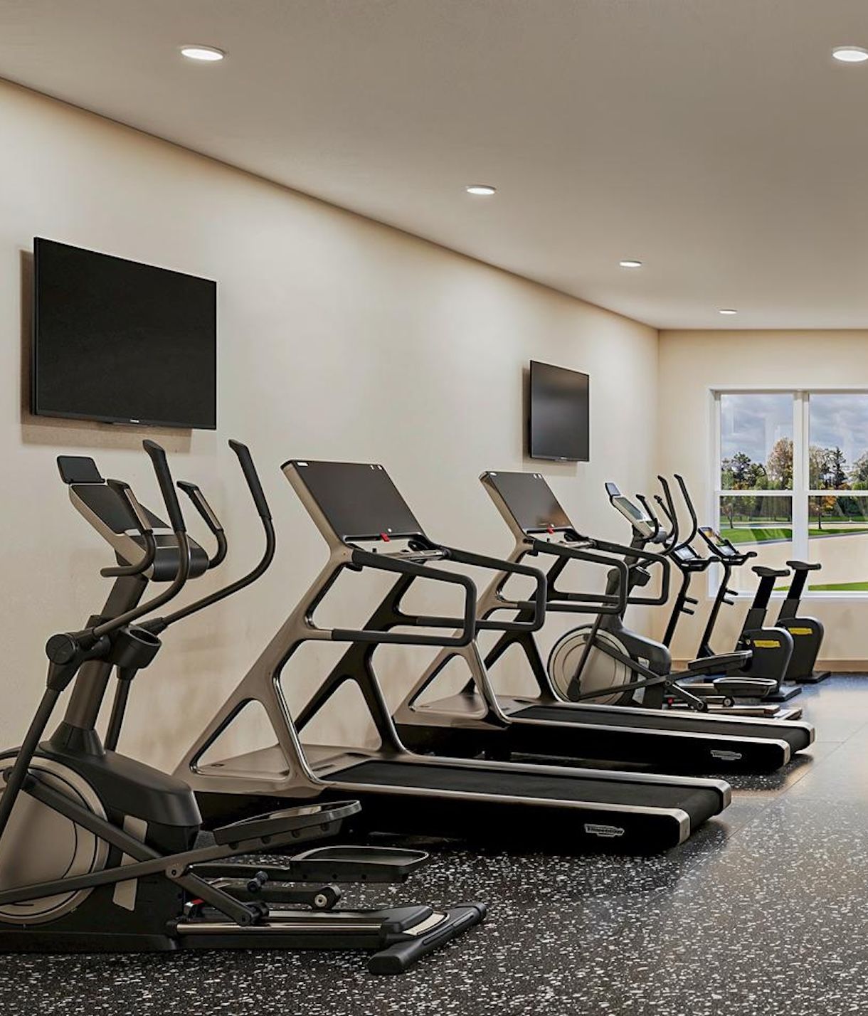Modern gym with treadmills, ellipticals, wall-mounted TVs, and large window showing outdoor scenery.
