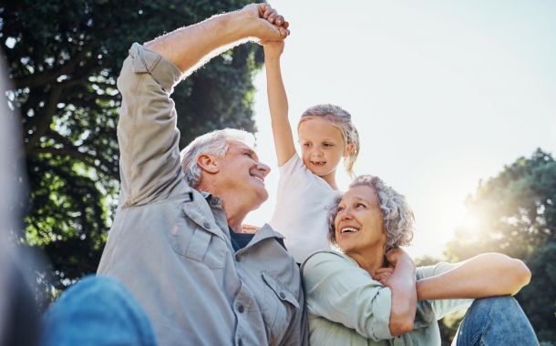Smiling grandparents and their granddaughter holding hands outdoors on a sunny day.